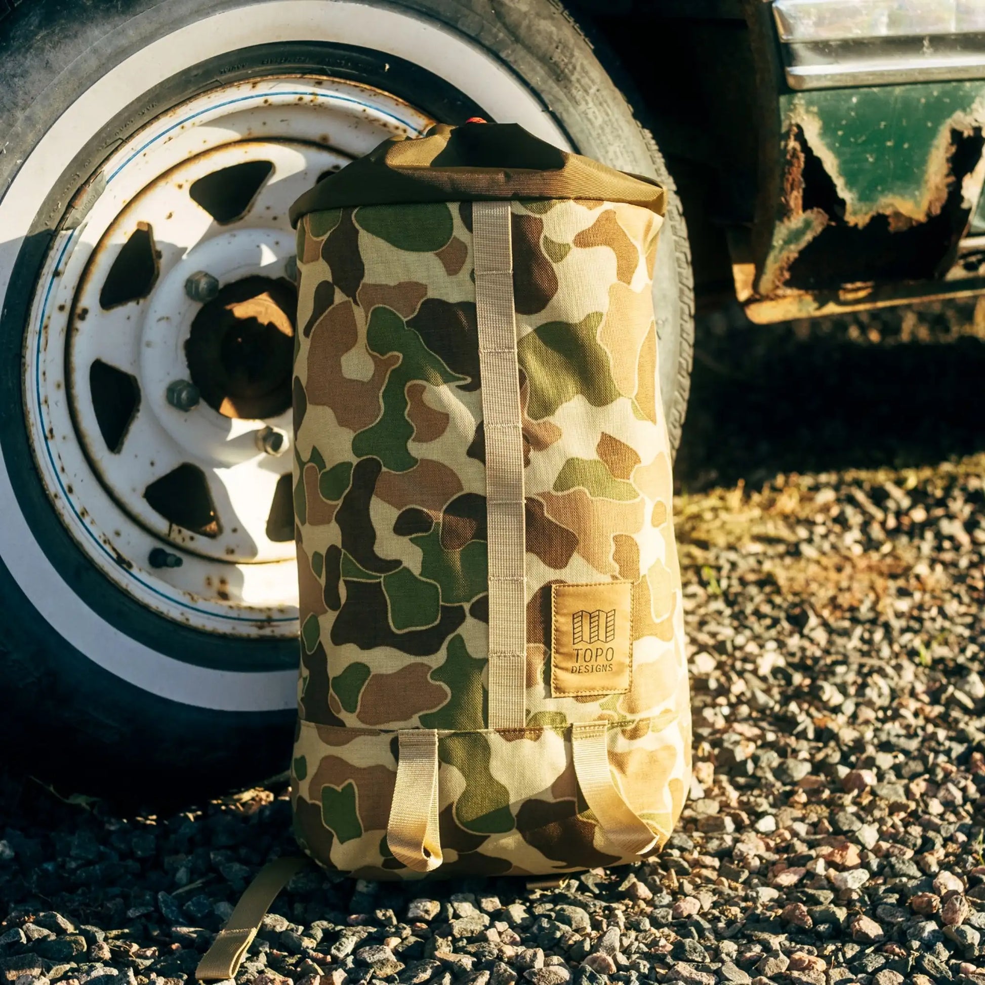 Duck camouflage backpack with tan and olive pattern, tan carry strap, and brand label visible on front.